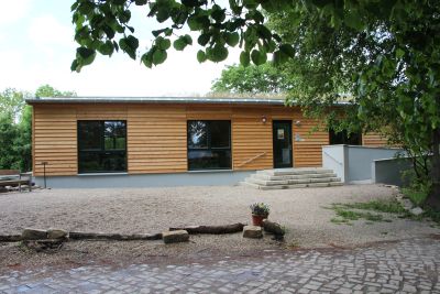 Vorderansicht des Schulungsgebäudes. Gebaut im Bungalowstil mit Holzverkleidung. Große Fenster, zum Eingang führen eine Treppe und eine Rollstuhlfaherrampe. Vorderansicht des Schulungsgebäudes. Gebaut im Bungalowstil mit Holzverkleidung. Große Fenster, zum Eingang führen eine Treppe und eine Rollstuhlfaherrampe.