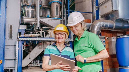 Das Bild zeigt zwei Personen mit einem Tablett vor einer Industrieanlage.