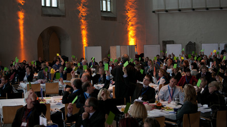 Das Bild zeigt die Teilnehmerinnen und Teilnehmer der Konferenz bei der Abstimmung zu einer der Publikumsfragen. Die Masse aller Beteiligten hält ein grünes Kärtchen in die Höhe.