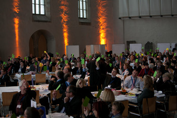 Das Bild zeigt die Teilnehmerinnen und Teilnehmer der Konferenz bei der Abstimmung zu einer der Publikumsfragen. Die Masse aller Beteiligten hält ein grünes Kärtchen in die Höhe.